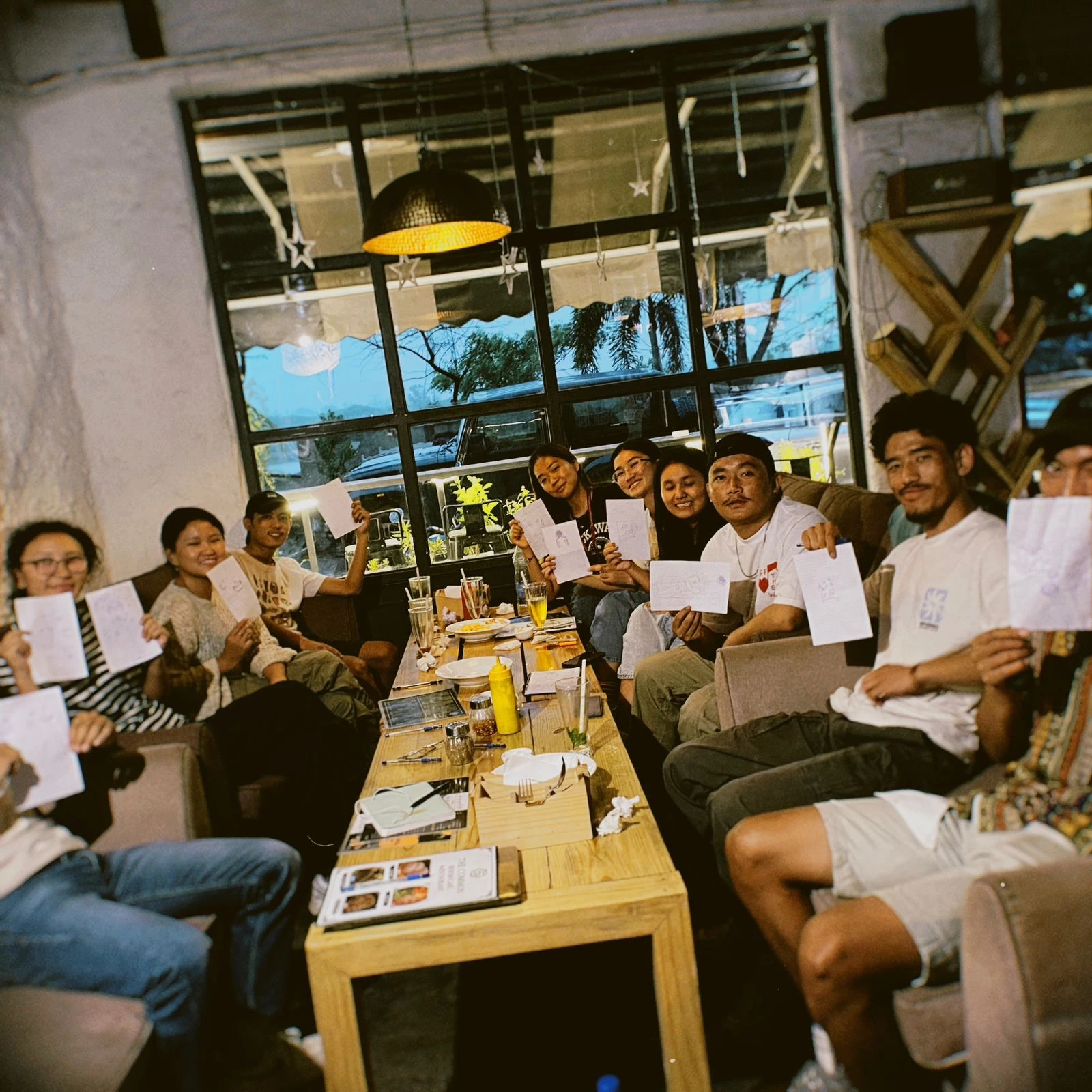 A group of Tibetan youth gathered around a café table, holding their reflection sketches at a Chisem community session.