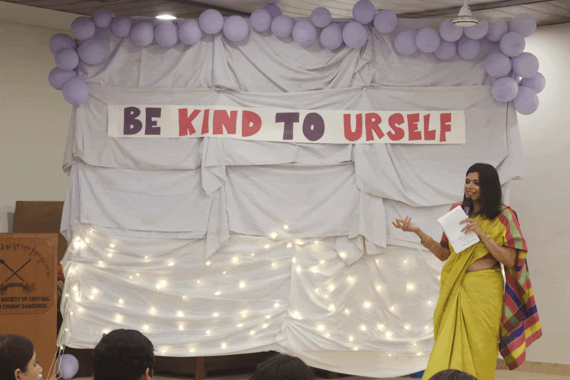 A speaker addresses youth at a Chisem “Be Kind to Urself” mental health event, framed by balloons and fairy lights.